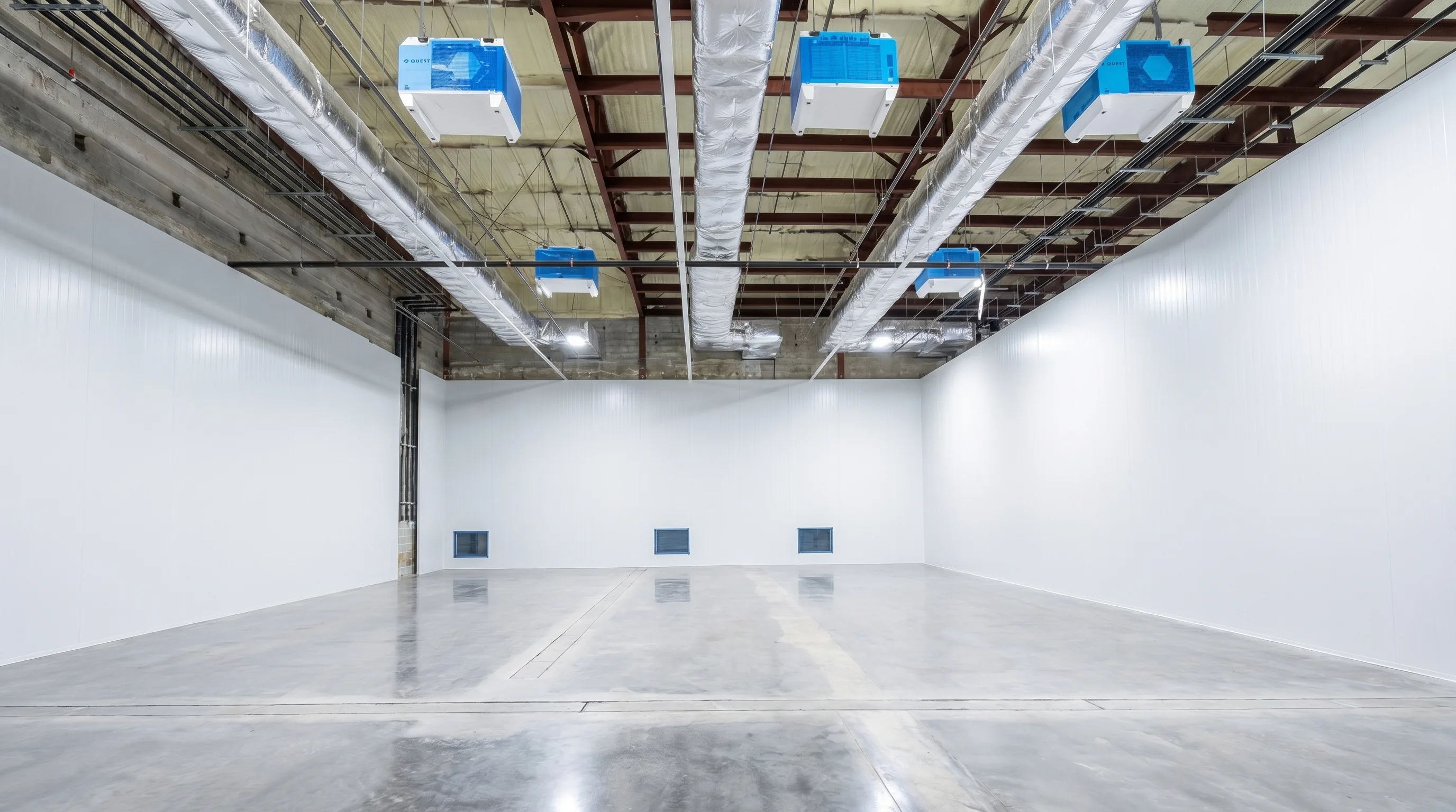 Empty cultivation room ready for fit-out: polished concrete floor, white walls, exposed steel-joist ceiling, six Quest dehumidifiers and insulated ductwork installed overhead — a healthcare-grade build before equipment goes in.