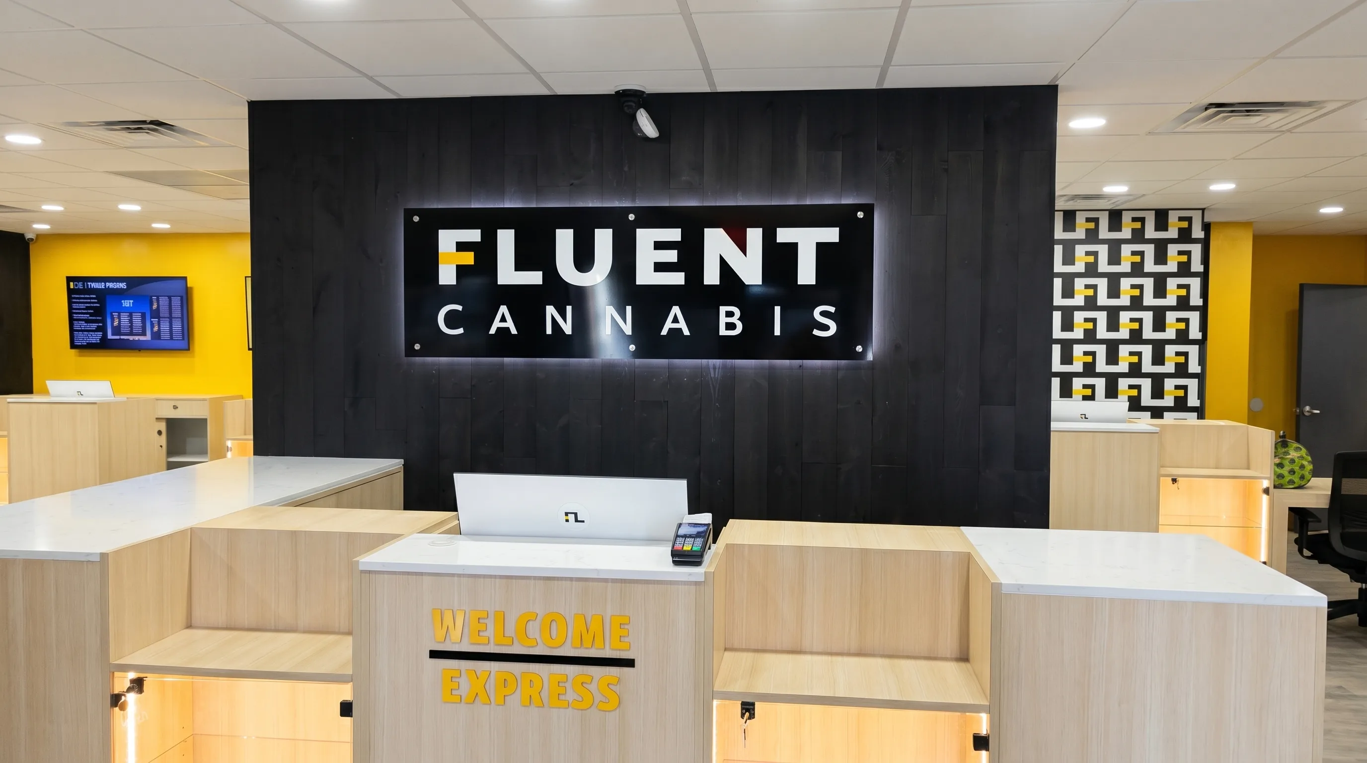 Interior of a Fluent Cannabis dispensary built out by Ronald Long: backlit FLUENT CANNABIS sign on dark wood paneling, wood reception counters, yellow Welcome Express counter — a Galleon construction portfolio buildout.